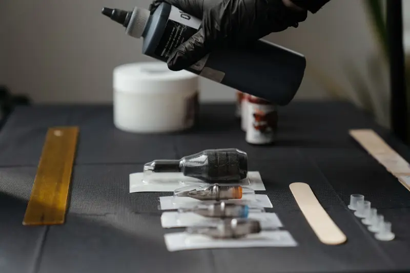 Tattoo machine, needle cartridges and a bottle laid out on a dark work table