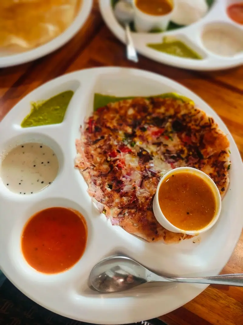 An onion uttapam on a white compartment plate with coconut chutney, tomato chutney and a cup of sambar
