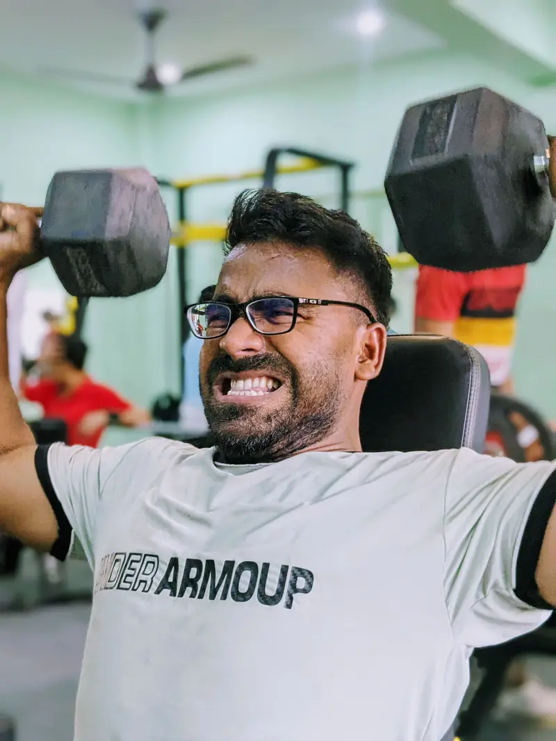 A man in glasses pressing two dumbbells overhead on a gym bench