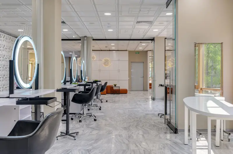 A bright salon with round lit mirrors, black styling chairs and a white marble floor.